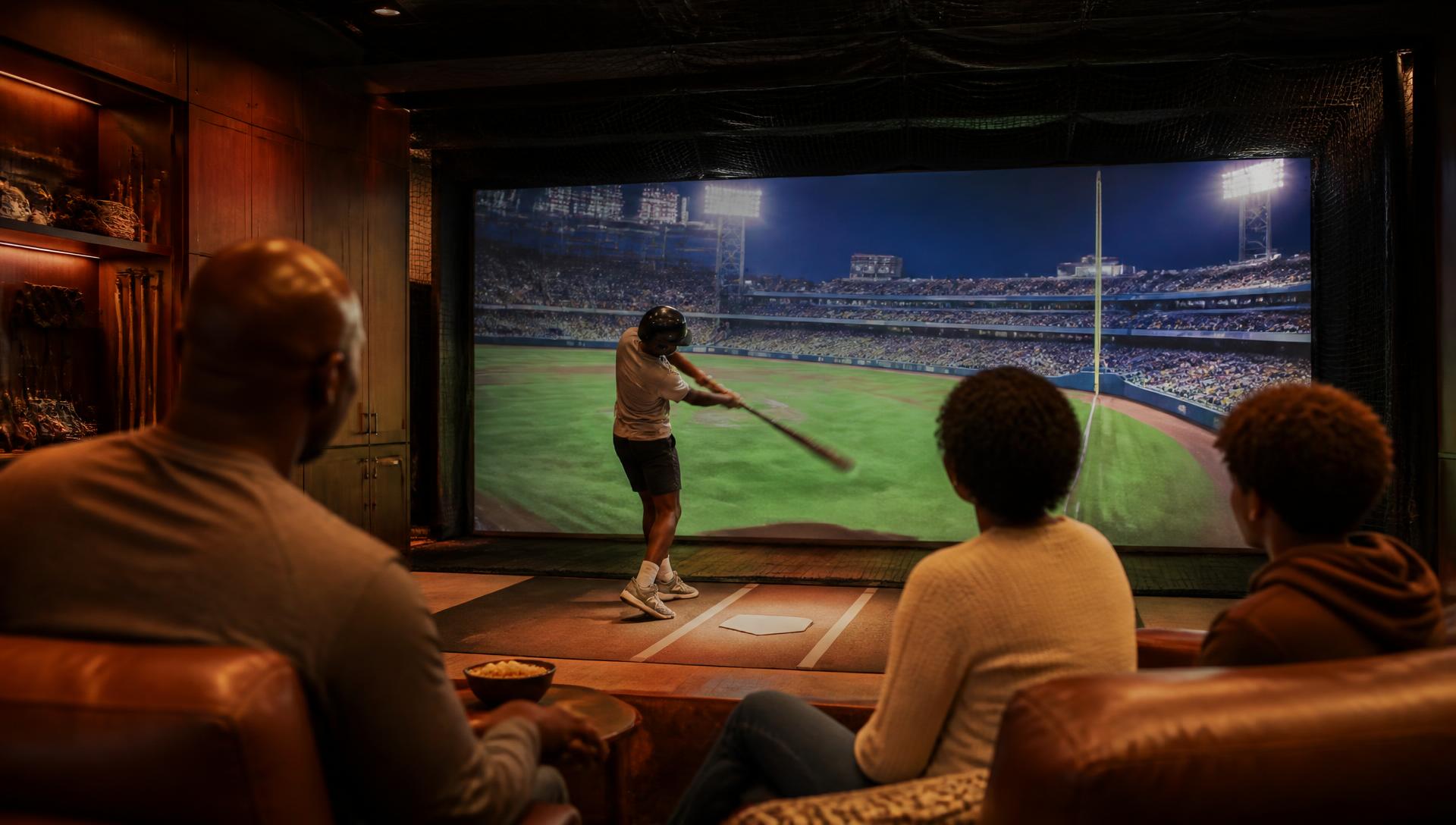 A big-league stadium filling the full-wall screen at Bat Club as a young hitter swings at home plate while his family watches from the walnut clubhouse lounge